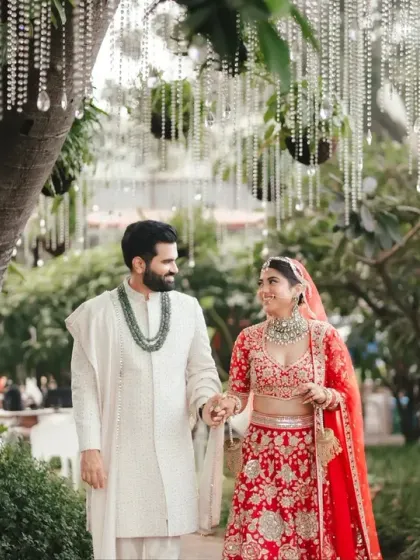 The couple walks hand-in-hand through a lush green pathway, sharing a loving glance. The contrast of her red lehenga and his white sherwani is visually stunning.