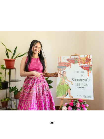 The bride poses next to a personalized welcome sign for her Mehendi ceremony, a lovely detail for the pre-wedding festivities.