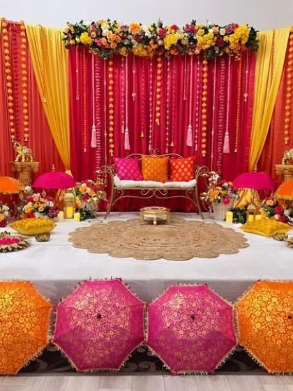 A stunning indoor Mehendi stage with red and yellow drapes. The decor is rich with traditional elements like embroidered umbrellas, brass elephants, and marigold flowers.