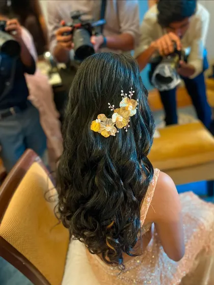 A behind-the-scenes shot of a soft, wavy half-updo being styled. The golden floral accessory adds a warm and elegant touch.