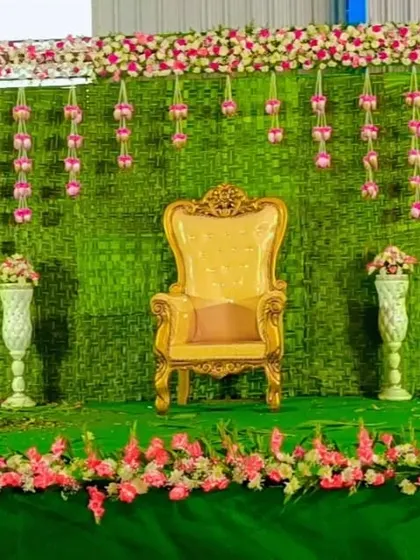 A simple yet elegant stage setup for a baby shower or seemantham. The green backdrop is made of woven leaves, with hanging pink flower garlands and a royal chair for the mother-to-be.