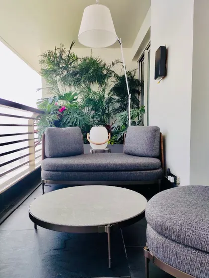 A cozy seating area on a small balcony. The design uses a comfortable daybed, a modern coffee table, and a large floor lamp, with a backdrop of dense tropical plants to create a private, room like feel.