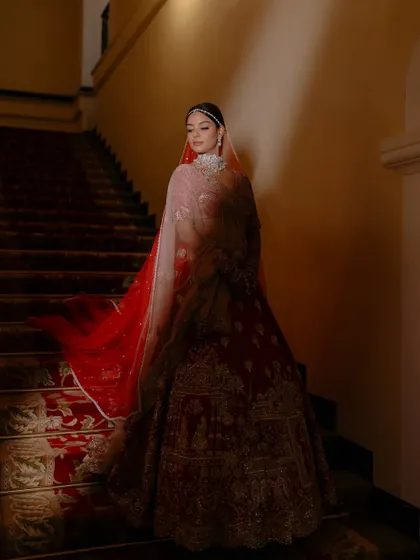 A dramatic and regal portrait on the stairs. The flowing red dupatta and the soft lighting create a truly cinematic bridal moment.