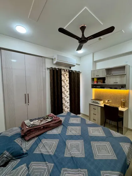 This bedroom includes a wardrobe with light-colored laminate and a compact study table with overhead cabinets and backlighting, making it a multi-functional space.