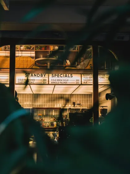 A creative shot looking into Stand By Coffee from the outside through foliage. This perspective creates a sense of discovery and highlights the warm, inviting glow of the cafe at night, making it feel like a hidden gem.