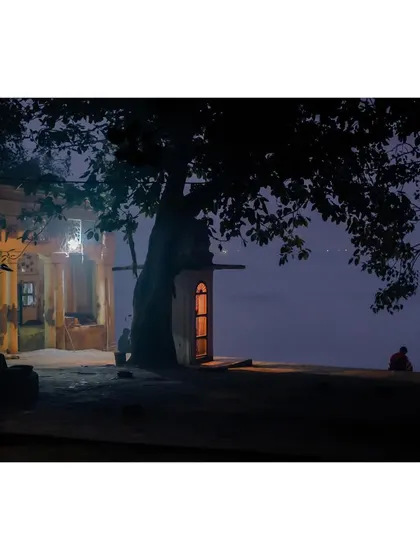A serene night scene on the banks of the Yamuna. A small, illuminated shrine sits under a large tree, while a lone figure sits by the water's edge in quiet contemplation.