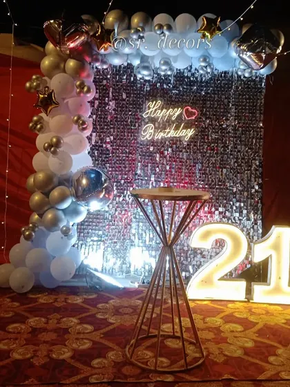 A dazzling 21st birthday setup in a banquet hall, using a silver shimmer wall, a white and silver balloon garland, and fairy lights for a magical effect.