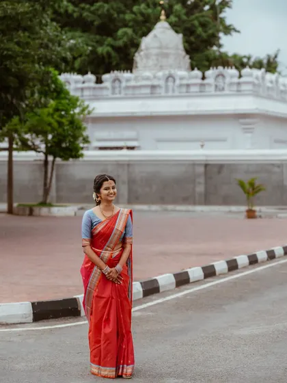 A wide shot showing the bride on her engagement day. The minimal makeup approach ensures her natural beauty is the focus, harmonizing perfectly with the traditional saree and temple setting.