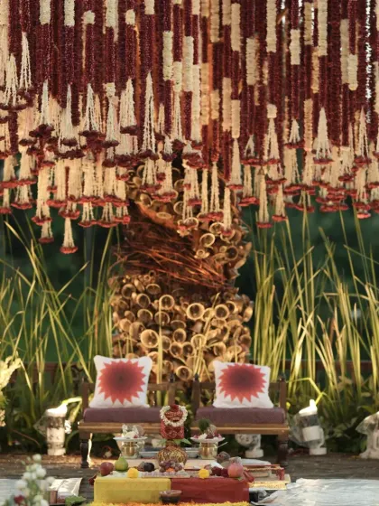 A closer look at the seating area within the Banaras Ghat mandap, with custom sun-motif cushions and a backdrop of wild grasses and pillars adorned with temple bells.