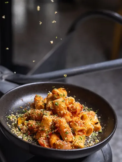 A sprinkle of herb crumbs falls onto a bowl of Rigatoni Arrabbiata. This adds a delightful crunch and an extra layer of flavor to the dish.