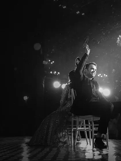 A dramatic black and white shot from a Sangeet performance. The stage lights, the rain or confetti, and the groom's pose create a powerful, rockstar-like image.