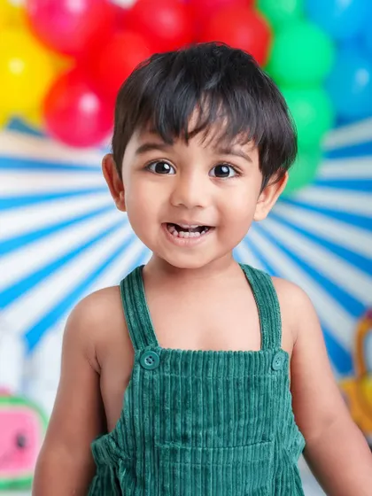 The brightest smile from this happy little guy. His green overalls and the colorful Cocomelon-inspired school bus background make for a cheerful and playful photo.