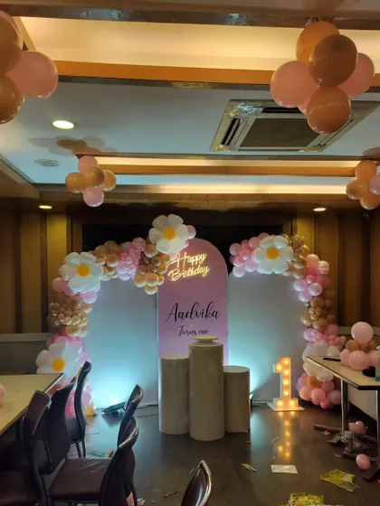 A sweet first birthday setup for Aadrika, with a pink and gold balloon arch, a personalized backdrop, and a light-up number '1'.