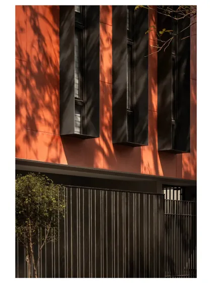 The deep red facade, inspired by traditional terracotta tones, has a tactile quality that interacts beautifully with light. Here, the shadows from the tree create a living pattern on the building's surface.
