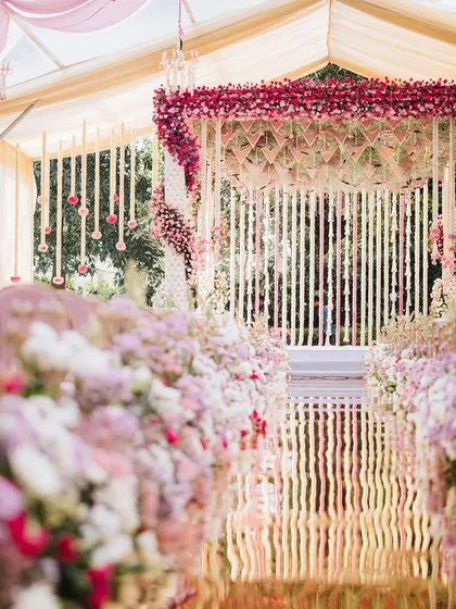 The aisle view from the mandap, looking back at the rows of pink chairs under a ceiling of draped fabric and chandeliers. This perspective highlights the symmetry and grandeur of the entire setup, designed for a truly cinematic experience.