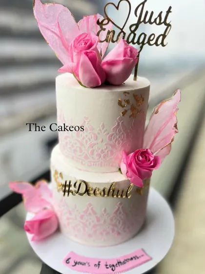 A romantic two-tier 'Just Engaged' cake in pink and white. It features an elegant stencil pattern, fresh pink roses, and delicate wafer paper wings, making it a beautiful centerpiece for an engagement party.