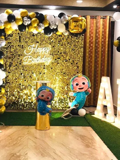 A Cocomelon-themed 4th birthday party set up at home. The decor features a gold shimmer wall, marquee letters, and character cutouts.