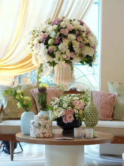A comfortable lounge area designed with elegant wooden furniture and soft cushions in pastel green and pink. Large floral arrangements add a touch of grandeur to the relaxed setting.