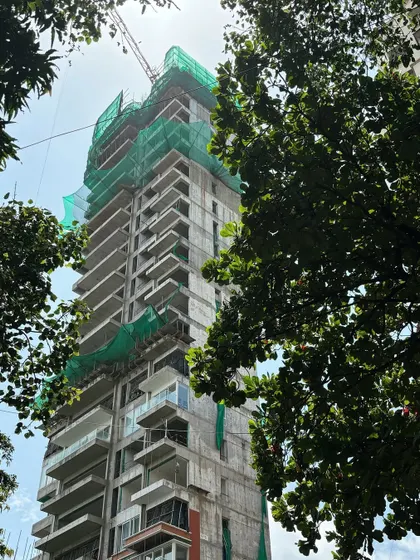 A view of a nearly completed luxury tower, framed by trees. This photo shows the building as it nears completion, with the protective netting coming down to reveal the finished façade and balconies.