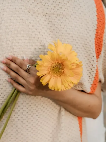 A creative and sweet detail shot. The bride-to-be holds a yellow flower behind her back, adding a touch of playful romance to their picnic-themed shoot.