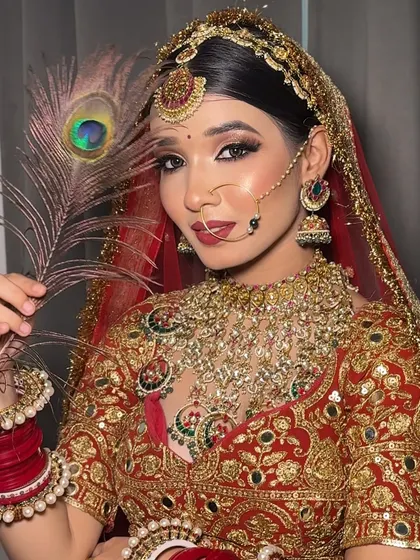 A regal portrait of a bride in a red lehenga, holding a peacock feather. Her makeup is classic and bold, with a focus on dramatic eyes and a red lip.