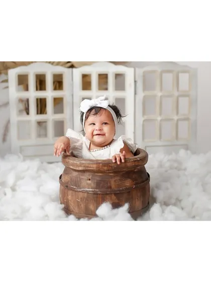 A smiling baby girl in a white dress, popping out of a wooden barrel prop in a dreamy cloud setup.