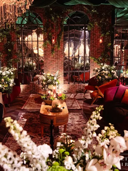 A lounge area in the glass house reception, featuring exposed brick walls, large arched windows with vintage mirrors, and lush white florals. The design was inspired by New York lofts and vintage London homes.