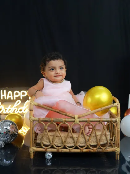A quiet moment of reflection for the birthday girl, sitting in a rustic wicker cot. The 'Happy Birthday' neon sign and golden balloons add a touch of modern elegance to her first birthday photoshoot.