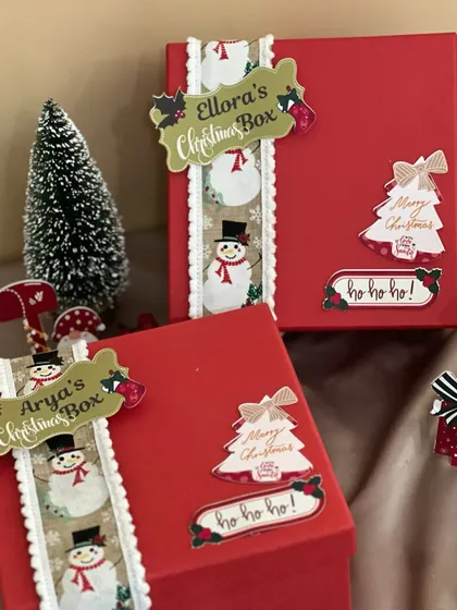 Making Christmas magical with personalized gift boxes. These bright red boxes are decorated with snowman ribbons and custom name tags for Ellora and Arya.