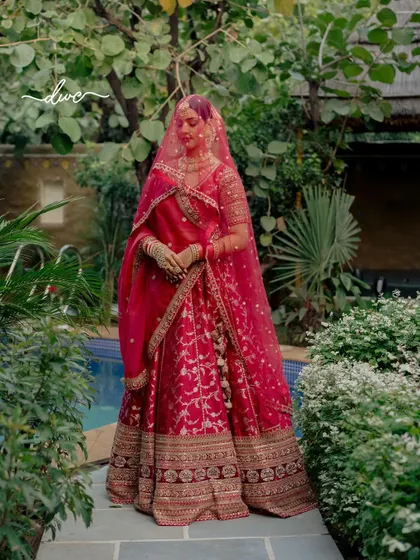 A full-length portrait of the bride in her vibrant red lehenga, standing amidst lush greenery, her veil creating a beautiful frame.