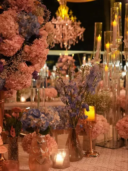 A detailed view of the layered tablescape, with tall glass candelabras rising behind lush arrangements of pink and blue flowers. The design creates a sense of depth and opulence.