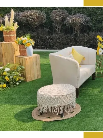 A chic and comfortable seating area in a garden setting. The white sofa and macrame stool are paired with wooden block tables topped with yellow floral arrangements, creating a perfect bohemian lounge.
