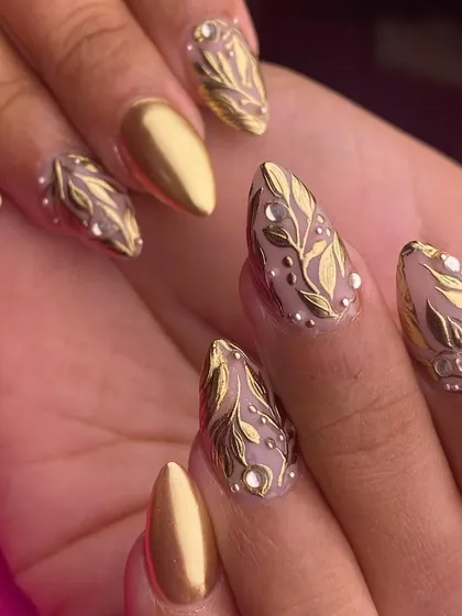 Pure opulence in a nail set. This design features matte gold chrome nails paired with accent nails showcasing intricate, raised golden leaf patterns over a sheer base, dotted with tiny crystals.