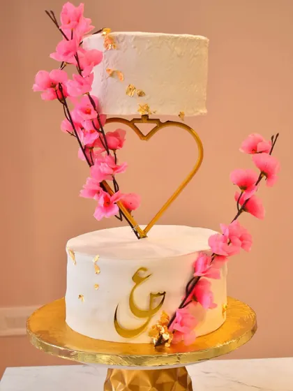 A modern and romantic floating engagement cake. This design uses a custom heart-shaped separator to create the illusion of a floating top tier, decorated with delicate pink cherry blossoms and personalized gold calligraphy.