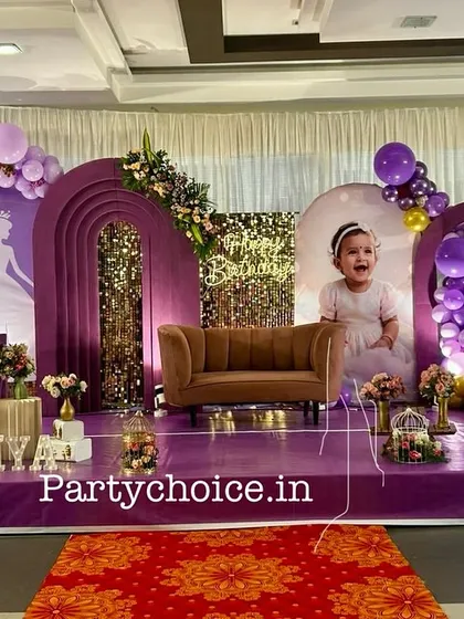 We designed this royal princess theme for a first birthday, using a rich purple color palette. The stage includes multiple arched backdrops, a miniature sofa for photos, a light-up number '1', and a princess carriage prop, all set on a purple carpeted stage.