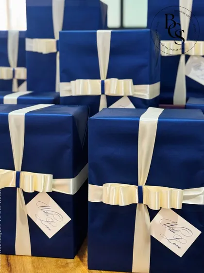 A stack of beautifully wrapped gifts in a deep royal blue. The classic wrapping is elevated by a contrasting cream-colored satin ribbon and a personalized "With Love" tag, ideal for a formal event or corporate gesture.