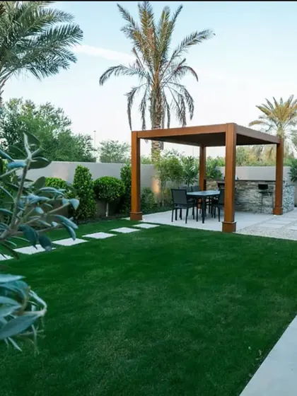 A completed backyard site transformation in Noida. This image shows a beautiful lawn with a wooden pergola for dining, surrounded by palm trees and other lush greenery.