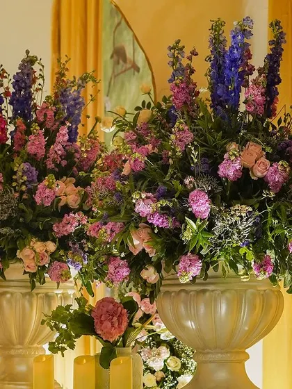 A close-up of two large floral arrangements in classic white urns, filled with a mix of pink, purple, and blue flowers.