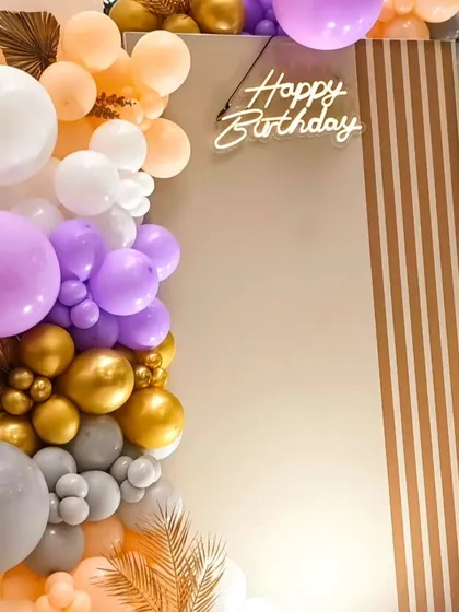 A close-up of a modern balloon garland with a 'Happy Birthday' neon sign. The mix of pastel colors with metallic gold and grey balloons creates a very chic look.