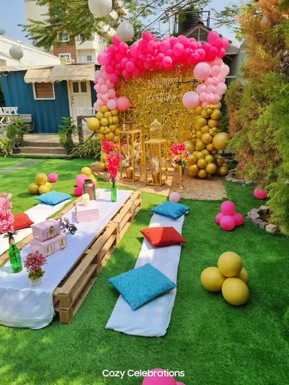A vibrant and fun outdoor party with a pink and gold theme. The setup includes a sequin wall, a large balloon arrangement, and low picnic tables for a relaxed party vibe.