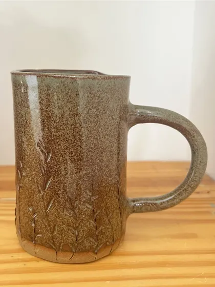 This tall, hand-built mug has a subtle texture from pressing plants into the clay. The earthy glaze gives it a rustic charm.