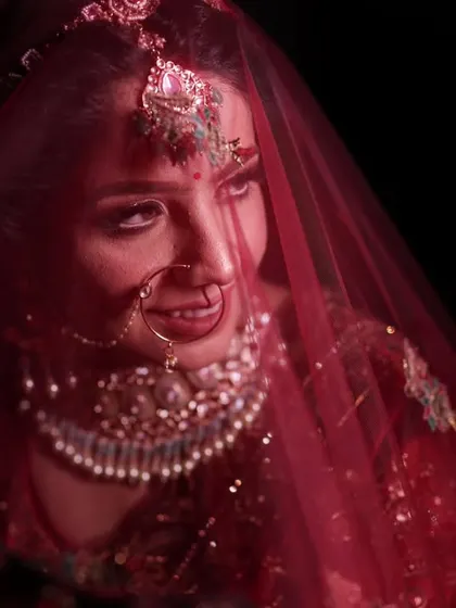 This is the style of bridal portrait I love to create. Using moody, dramatic lighting, I focus entirely on the bride, capturing that quiet moment of anticipation under the red veil. The rich colors of her lehenga and the intricate details of her jewelry, like the nath and maang tikka, stand out beautifully against the dark background.