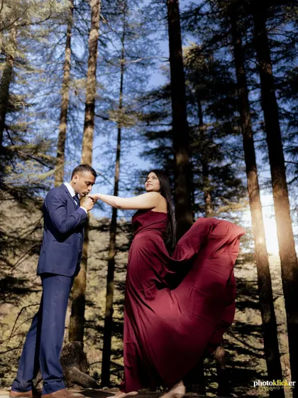 A classic romantic gesture as the man kisses the woman's hand, her flowing maroon gown contrasting beautifully with the tall pine trees.