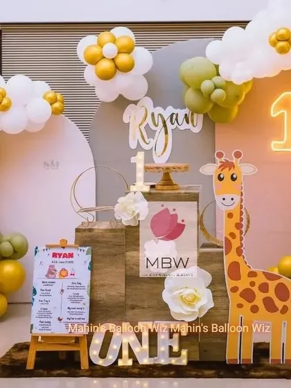 A cheerful and modern first birthday setup with a friendly giraffe character. The design uses a warm color palette of olive green, orange, and gold, complemented by a neon "1" and a personalized milestone board.
