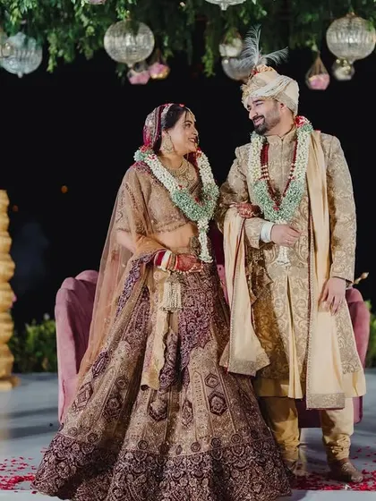 A portrait of the couple on their wedding day, standing on a stage adorned with golden pots and a canopy of hanging greenery.