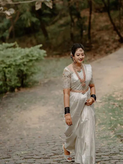 A bride in a stunning white saree walks along a paved path, showcasing her full bridal attire in a natural, outdoor setting.