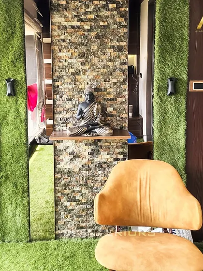 A tranquil corner featuring a stone-clad accent wall with a small shelf for a Buddha statue. The wall is flanked by panels of artificial grass, bringing an element of nature indoors.