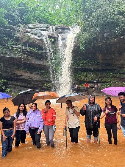 A group of friends with umbrellas standing in the reddish-brown water of a waterfall pool. The colours of the monsoon are truly unique.