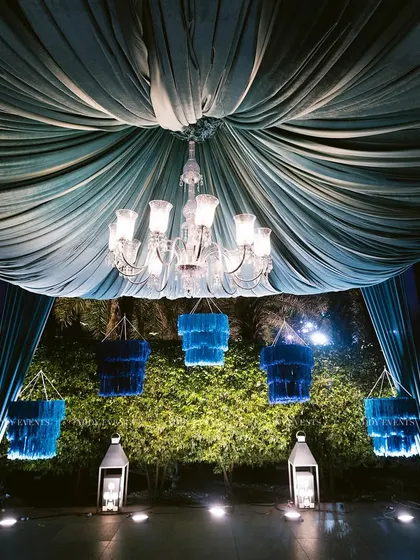 This welcome lounge setup features a dramatic royal blue draped canopy with a central crystal chandelier. The hanging blue tassel lanterns and floor lanterns add layers of light and color, creating a rich and inviting ambiance.