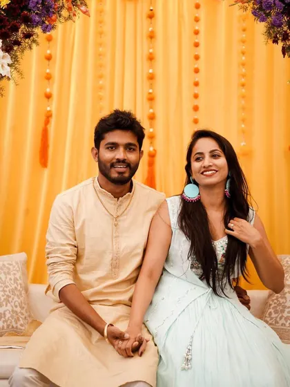A happy couple at their Mehendi ceremony. The decor features a bright yellow backdrop with orange tassels and a beautiful multi-colored floral arch.